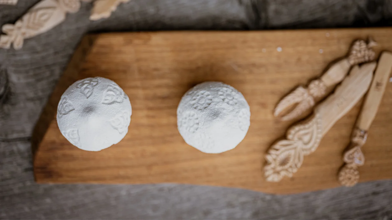 Two round cheeses with patterns and carved wooden utensils on a wooden board.