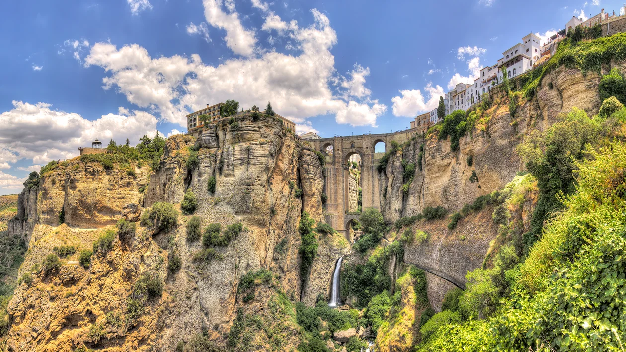 Stone bridge spans a deep gorge with cliffs, houses on top, and greenery below under a bright sky.