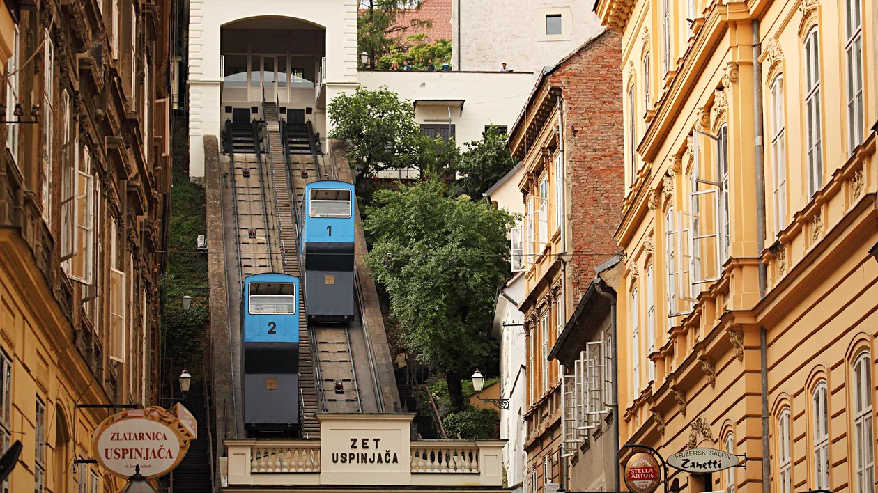 Two blue funicular cars move up and down a steep track between old city buildings.