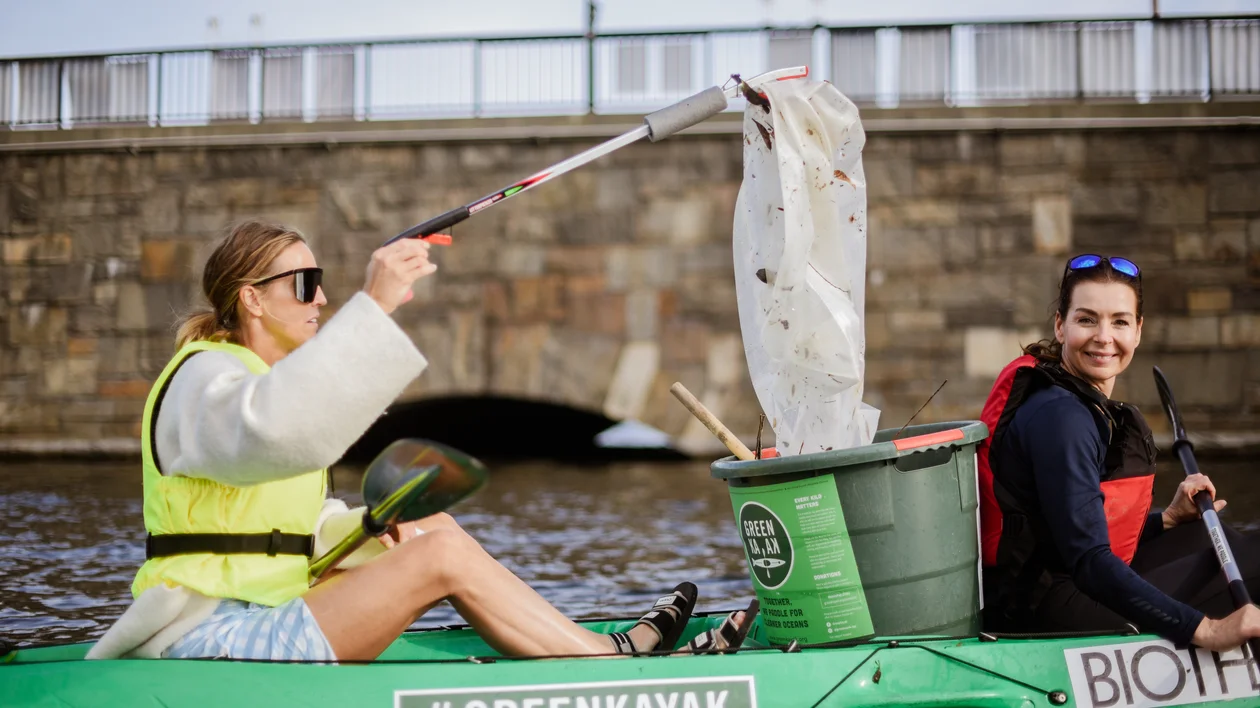Two people in a green kayak collect trash from the water near a stone bridge.