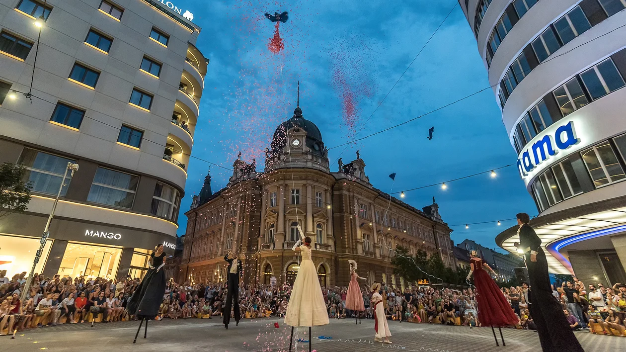 Street performance with people wearing long dresses, confetti falling, crowd watching at dusk.