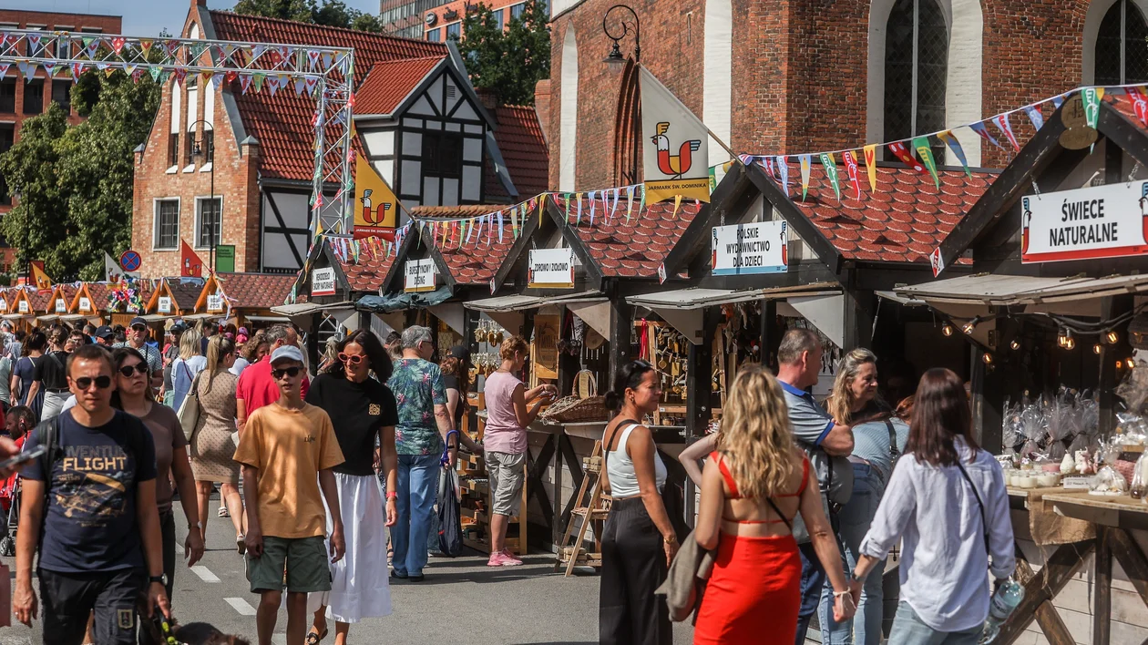Crowds walk among outdoor market stalls with colorful flags and historic buildings.