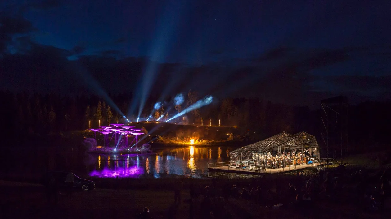 Outdoor concert at night with stage lights reflecting on a lake and beams lighting the sky.