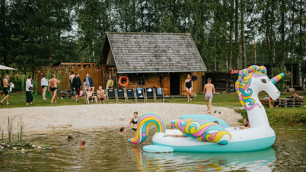 People swimming and relaxing by a pond with a large unicorn float and a wooden cabin nearby.