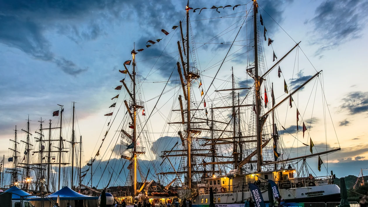 Tall sailing ships docked at sunset, with flags and tents nearby.