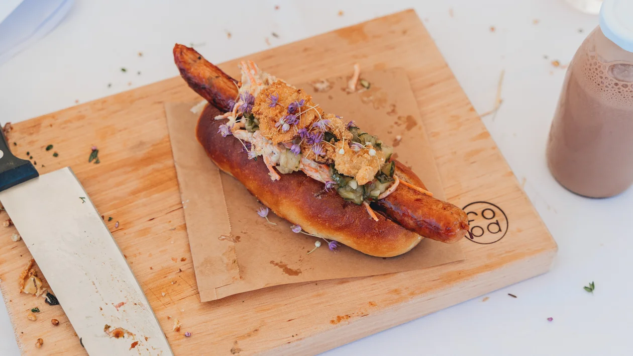 Gourmet sausage in a bun topped with slaw on a wooden board, knife beside it.