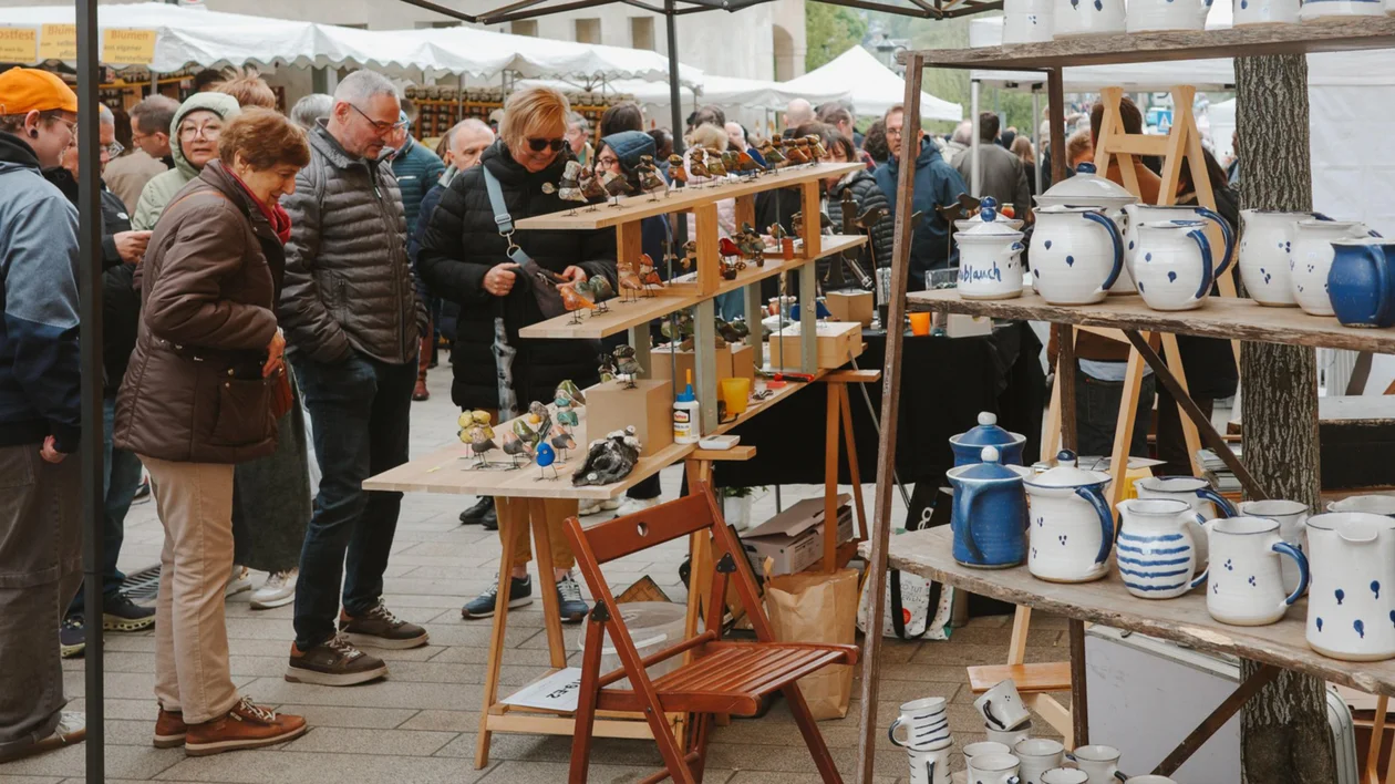 People browsing ceramic pottery at an outdoor market, shelves filled with mugs and jugs.