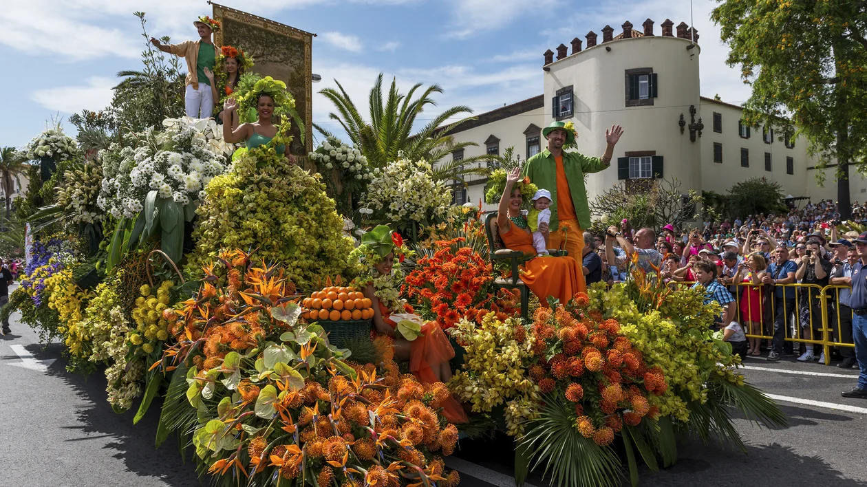 Colorful parade float covered in flowers passes a cheering crowd on a sunny day.