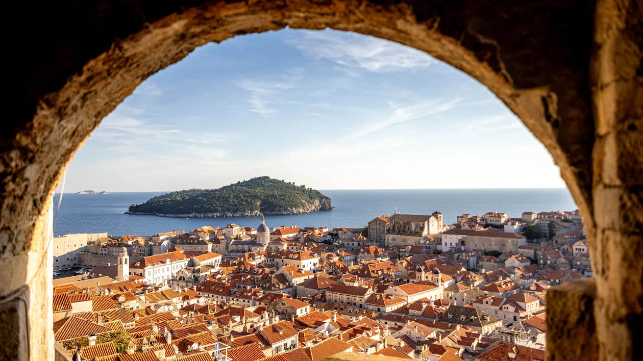 View of orange rooftops and the sea through a stone archway, with a green island in the distance.