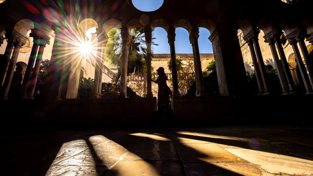Sunlight streaming through arches in a stone courtyard with a silhouetted statue.
