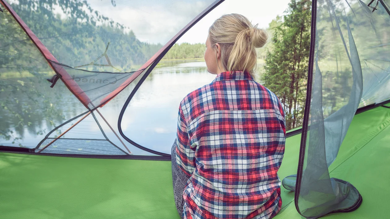 Woman in plaid shirt sitting in a tent, looking out at a lake and trees.