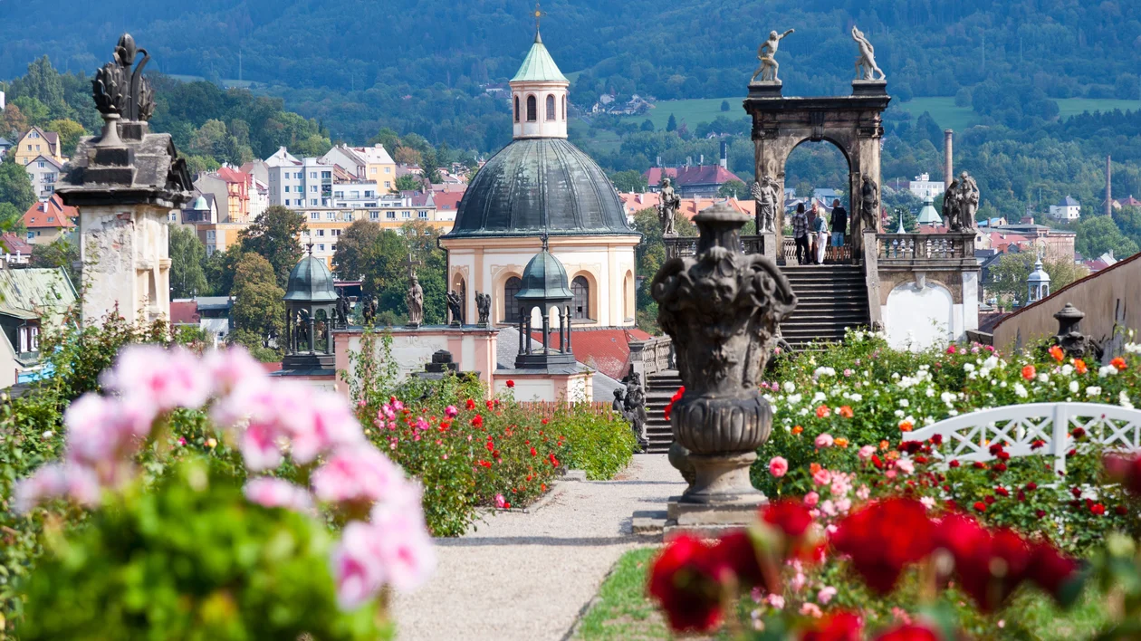 Colorful flower garden with statues, overlooking a dome building and a scenic town backdrop.