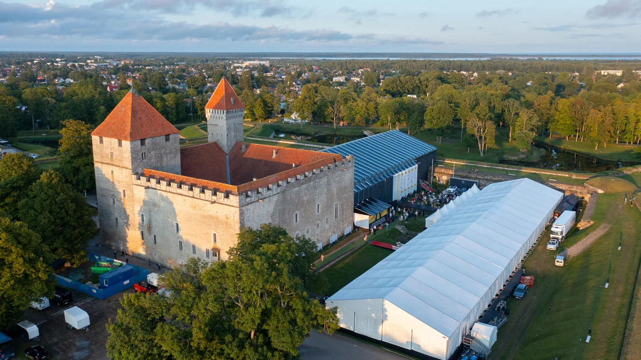 Medieval stone castle with red roofs beside large white tents, surrounded by greenery.