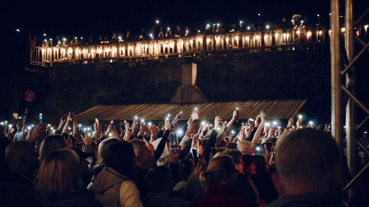 Crowd at night holding up lit phones, with bridge lights shining above.