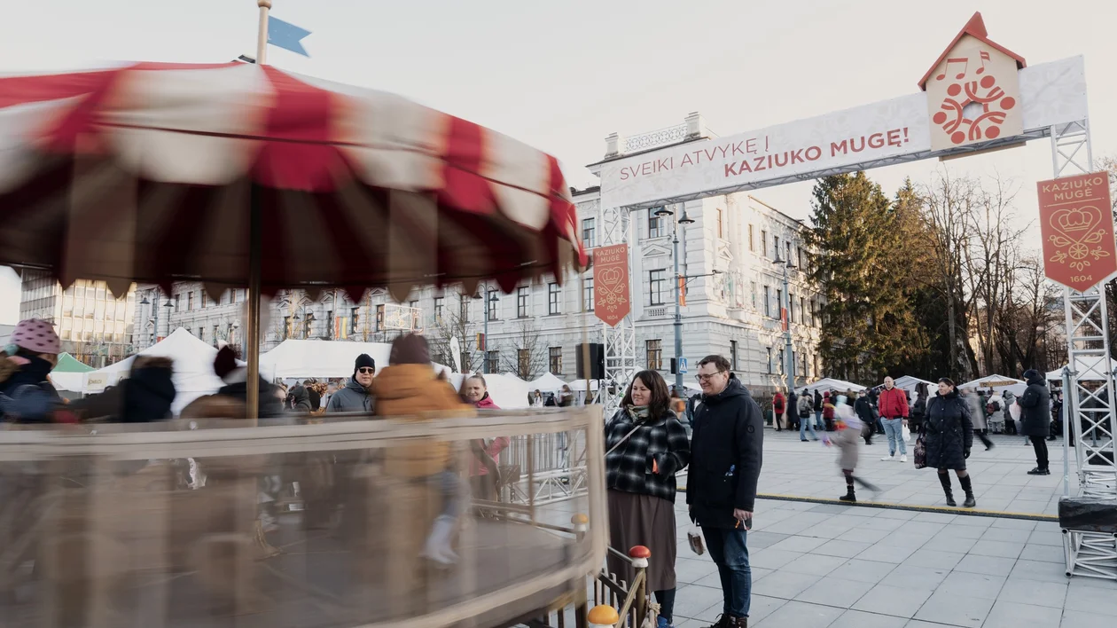 People gather at an outdoor market near a spinning carousel and festive entrance arch.