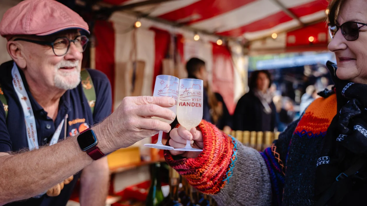 Two people clinking glasses and smiling at an outdoor market stall.
