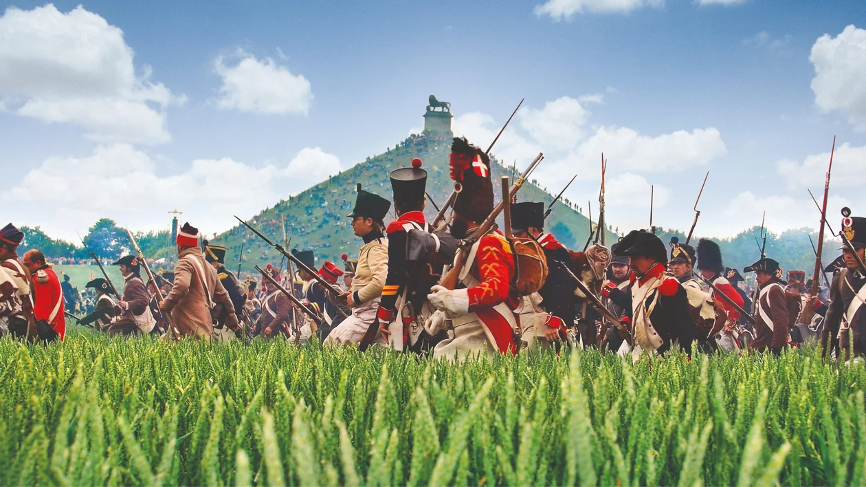 Soldiers in historical uniforms march through a grassy field under a blue sky with scattered clouds.