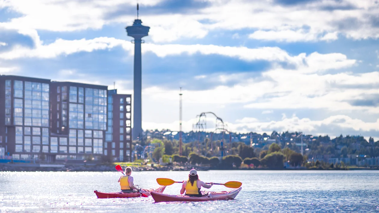 Two people kayaking on a lake with city buildings and a tower in the background.