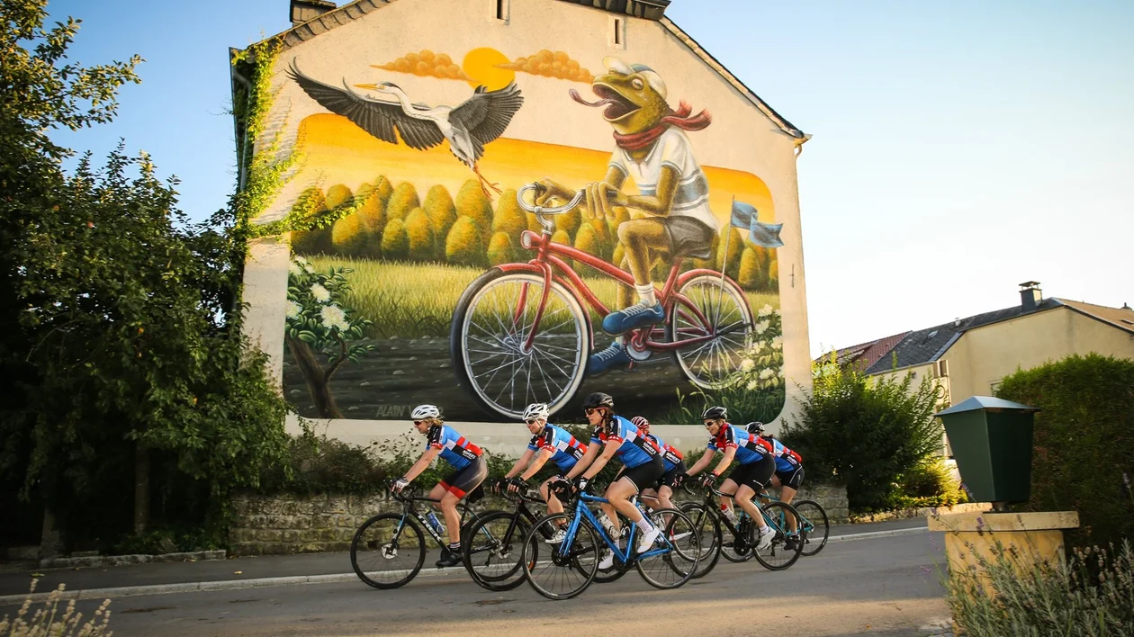 Cyclists ride past a mural of a frog on a bicycle with a stork, set on a sunny street.