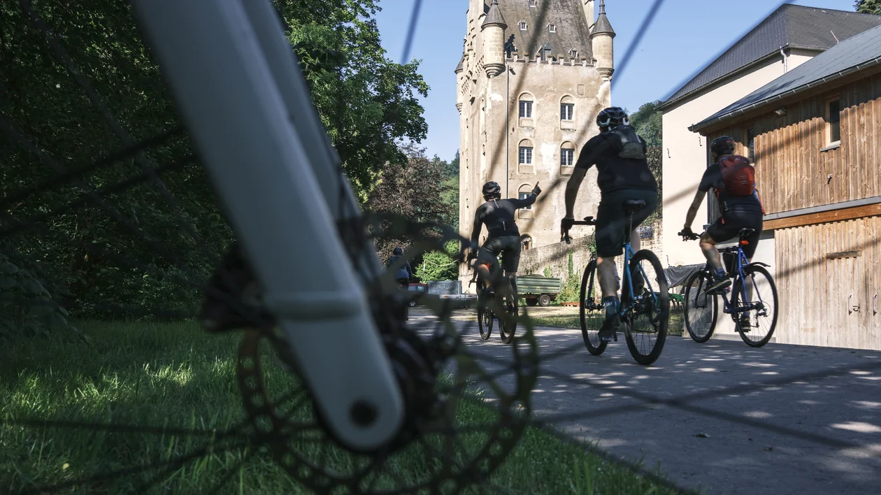 Three cyclists ride past a castle, framed by a close-up of a bicycle wheel.