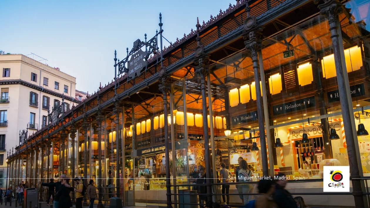 Glass-walled market building glowing with warm lights at dusk, people gathered outside.