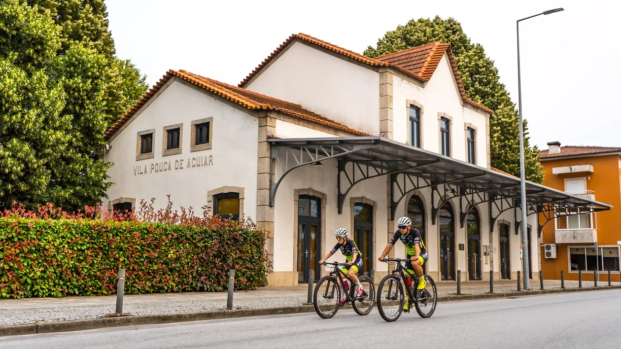 Two cyclists ride past a white building with a red roof and green trees.