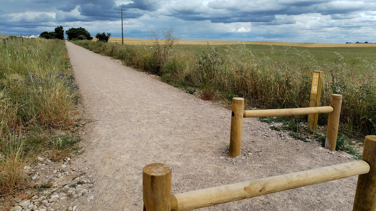 Gravel path with wooden barrier, grassy fields, and cloudy sky.