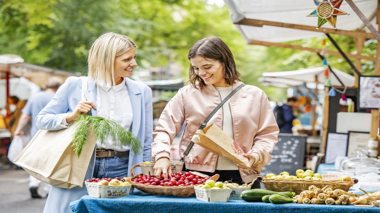 Two women shopping for produce at an outdoor market, smiling and chatting.