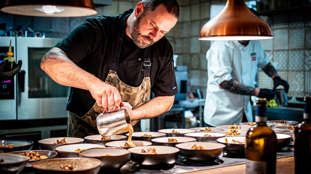 Chef pouring sauce onto plated dishes in a professional kitchen under warm lights.