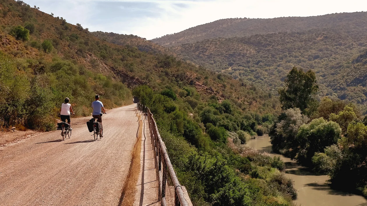 Two cyclists ride on a dirt road beside a river through green hills.