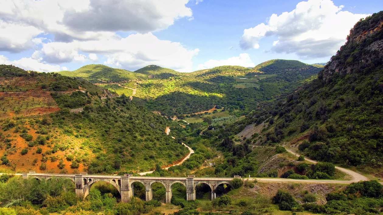 Stone railway bridge crosses a lush green valley with mountains under a blue sky with clouds.