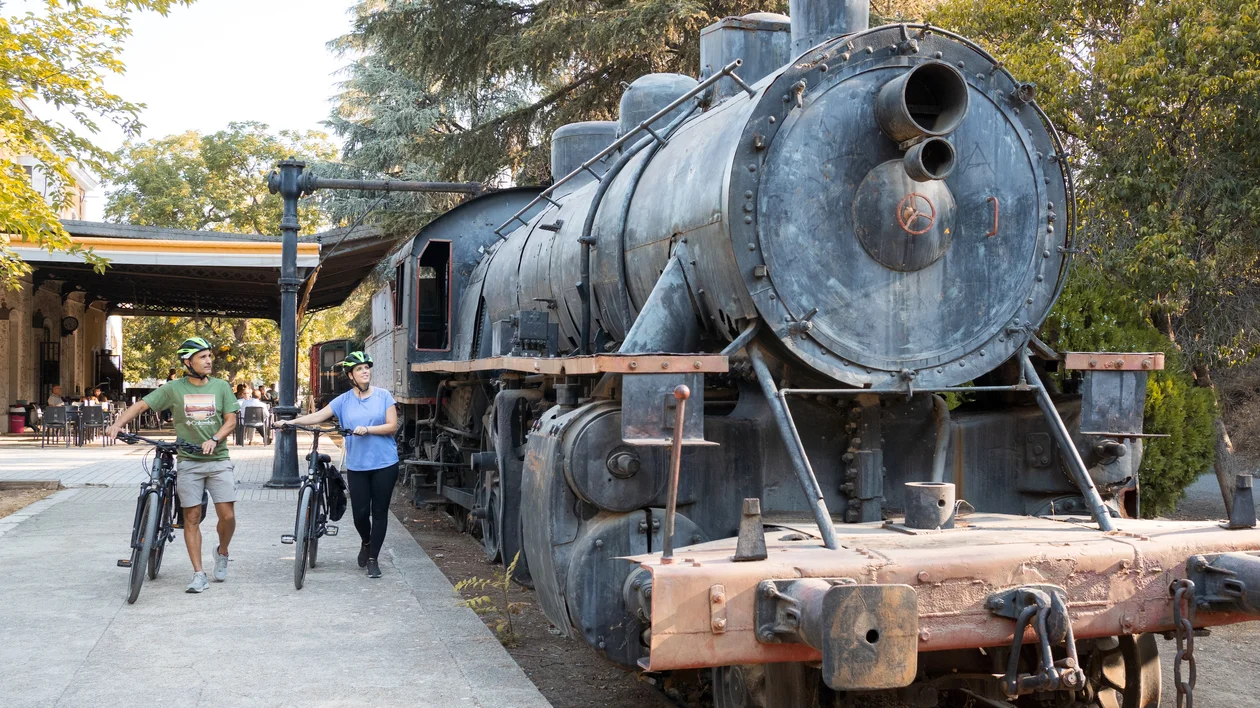 Two people with bicycles walk past an old black steam locomotive outdoors.