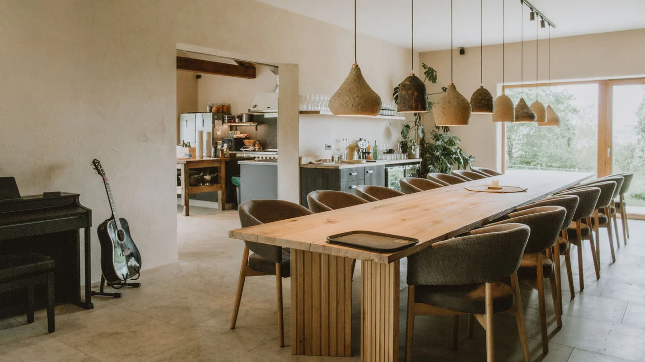 Long wooden dining table with chairs, pendant lights, and bright open kitchen in background.