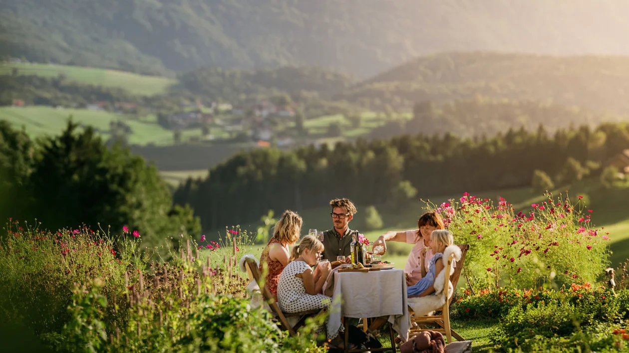 Four people enjoying a meal at a table in a scenic, sunlit mountain meadow.