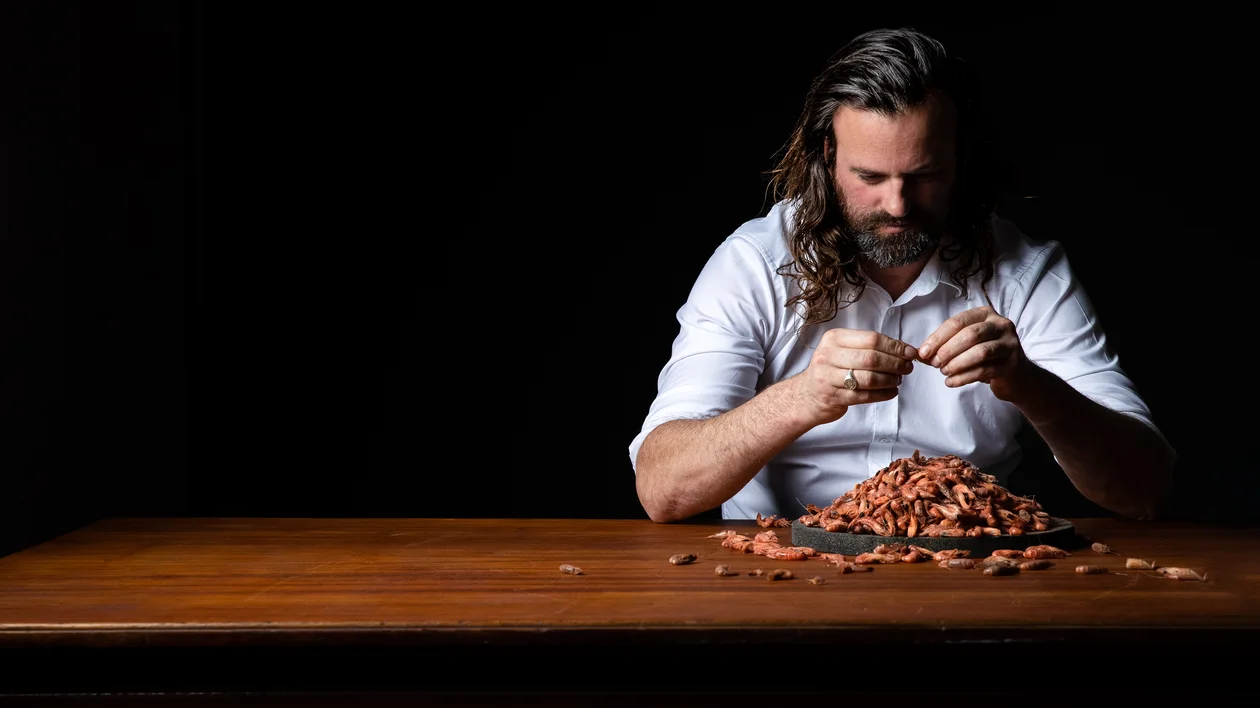 Man in a white shirt shelling almonds at a wooden table against a dark background.