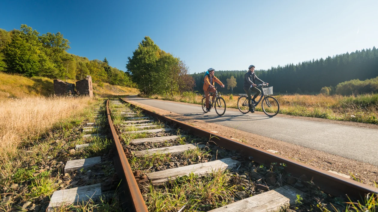 Two people biking on a path next to old railroad tracks under a clear blue sky.