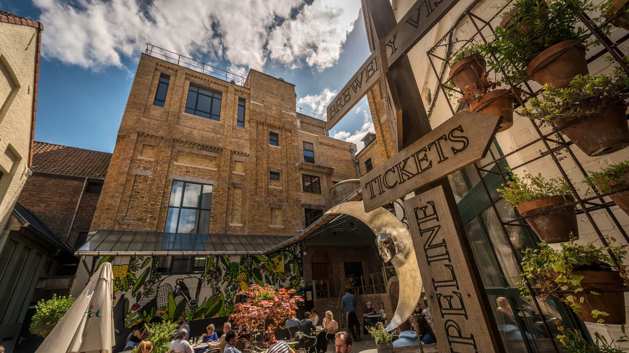 Sunny courtyard with people sitting, potted plants, and directional signs near brick buildings.