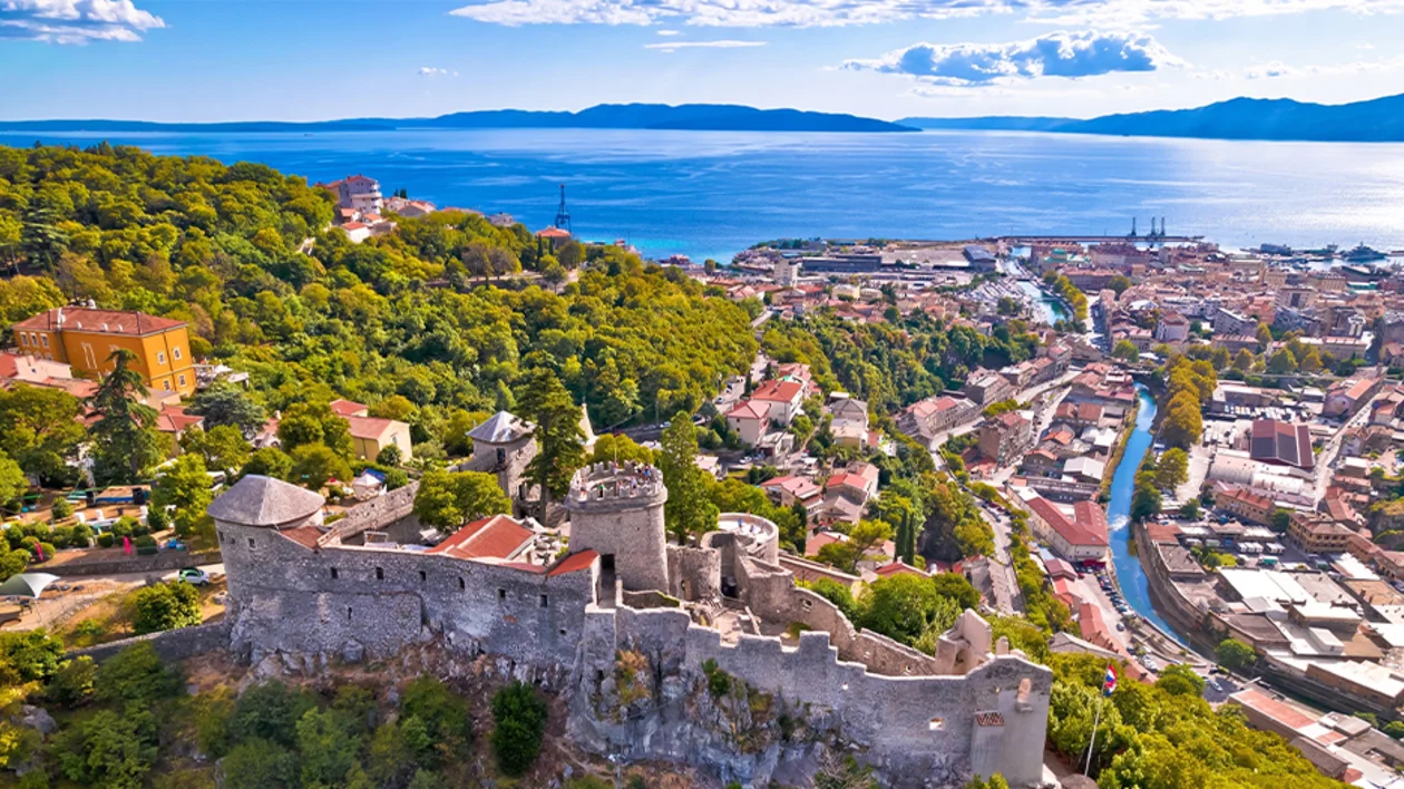 Hilltop stone fortress overlooking a coastal city with blue sea and green hills.