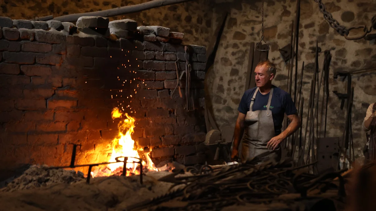 Blacksmith standing by a glowing forge in a rustic workshop with tools on the wall.
