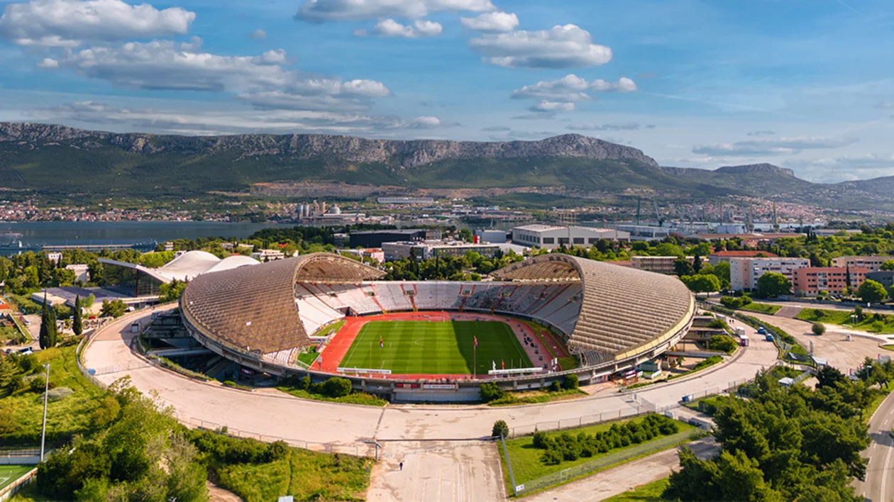 Large open-air stadium with a running track, surrounded by city and mountains.