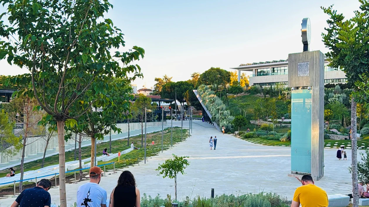 People sitting on grass near a modern park with trees, pathway, and buildings in the background.