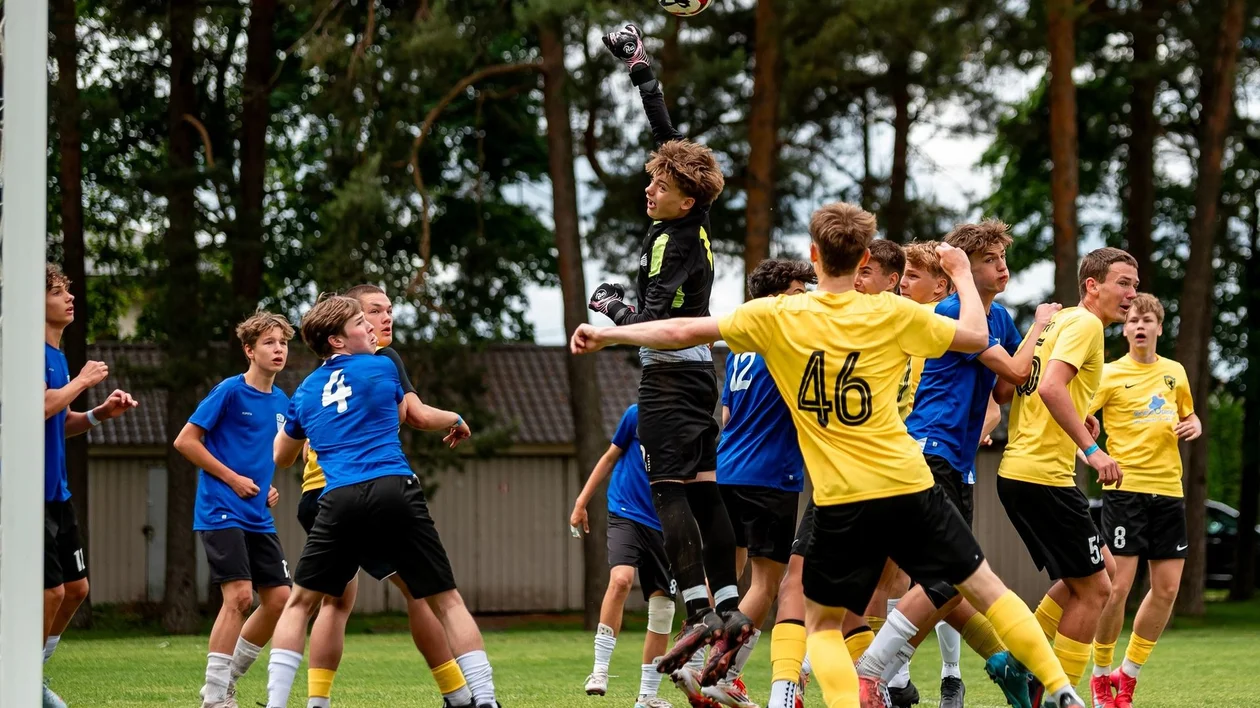 Goalkeeper jumps to catch soccer ball as players in blue and yellow crowd the goal area.