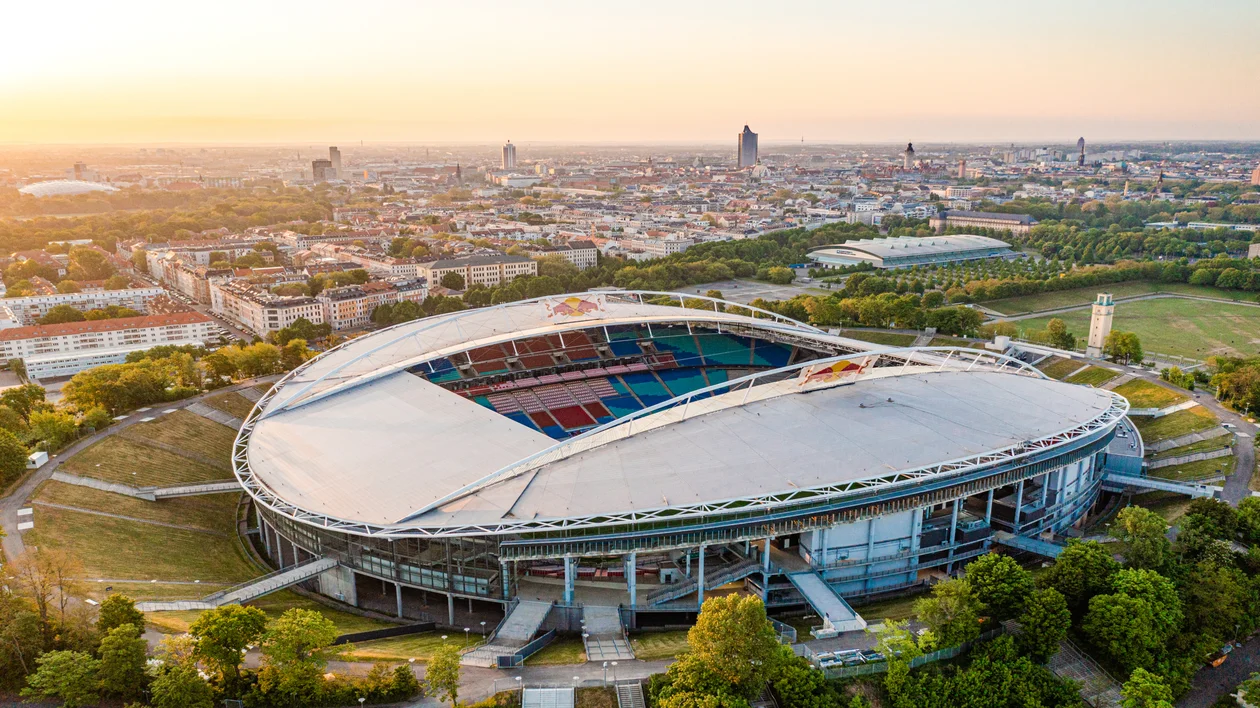 Large modern stadium surrounded by trees, cityscape in the background at sunset.