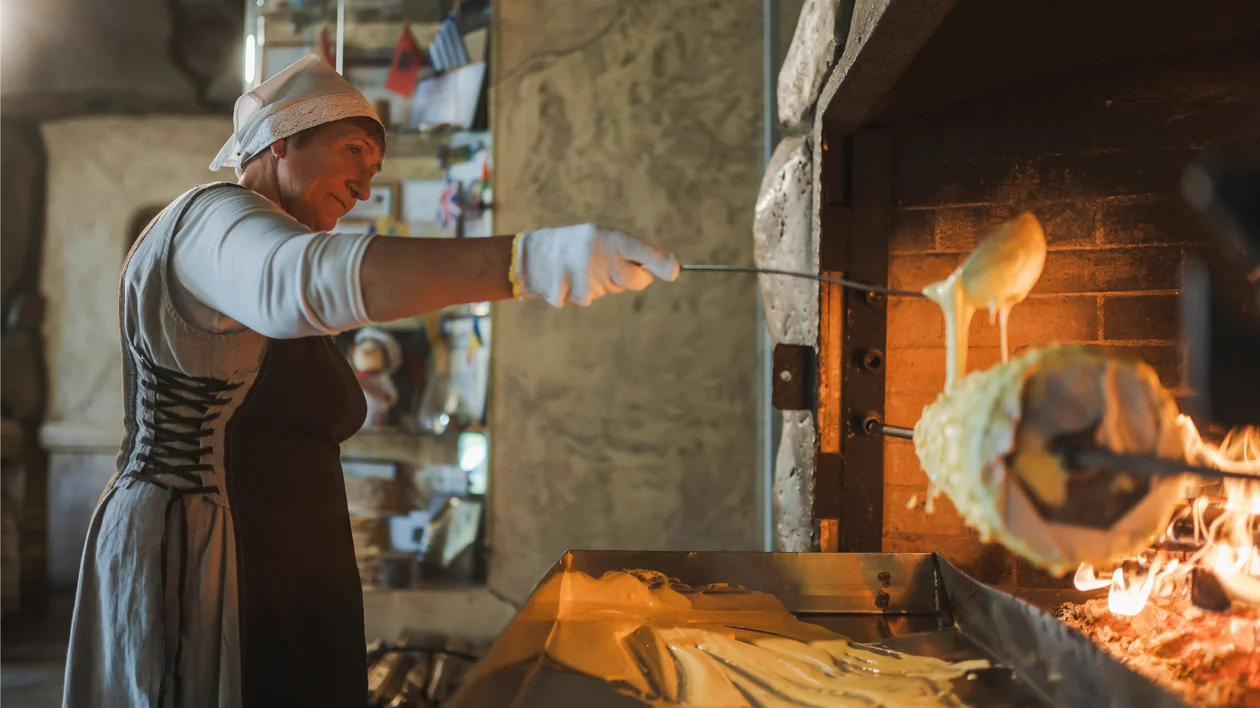 Woman baking a large cake on a spit over an open fire in a rustic kitchen.