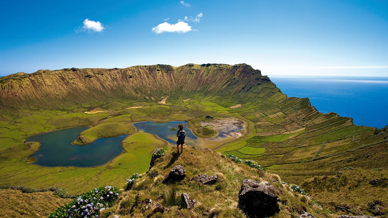 Green volcanic crater with lakes under a clear blue sky and ocean in the background.