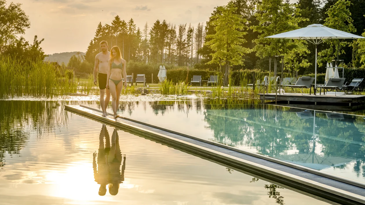 Couple walking beside a pool at sunset with trees and lounge chairs in the background.