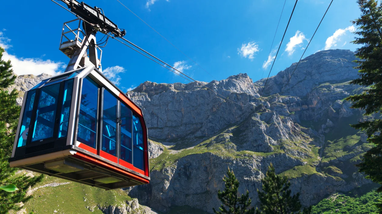 Blue cable car ascending over green trees with rocky mountains in the background.