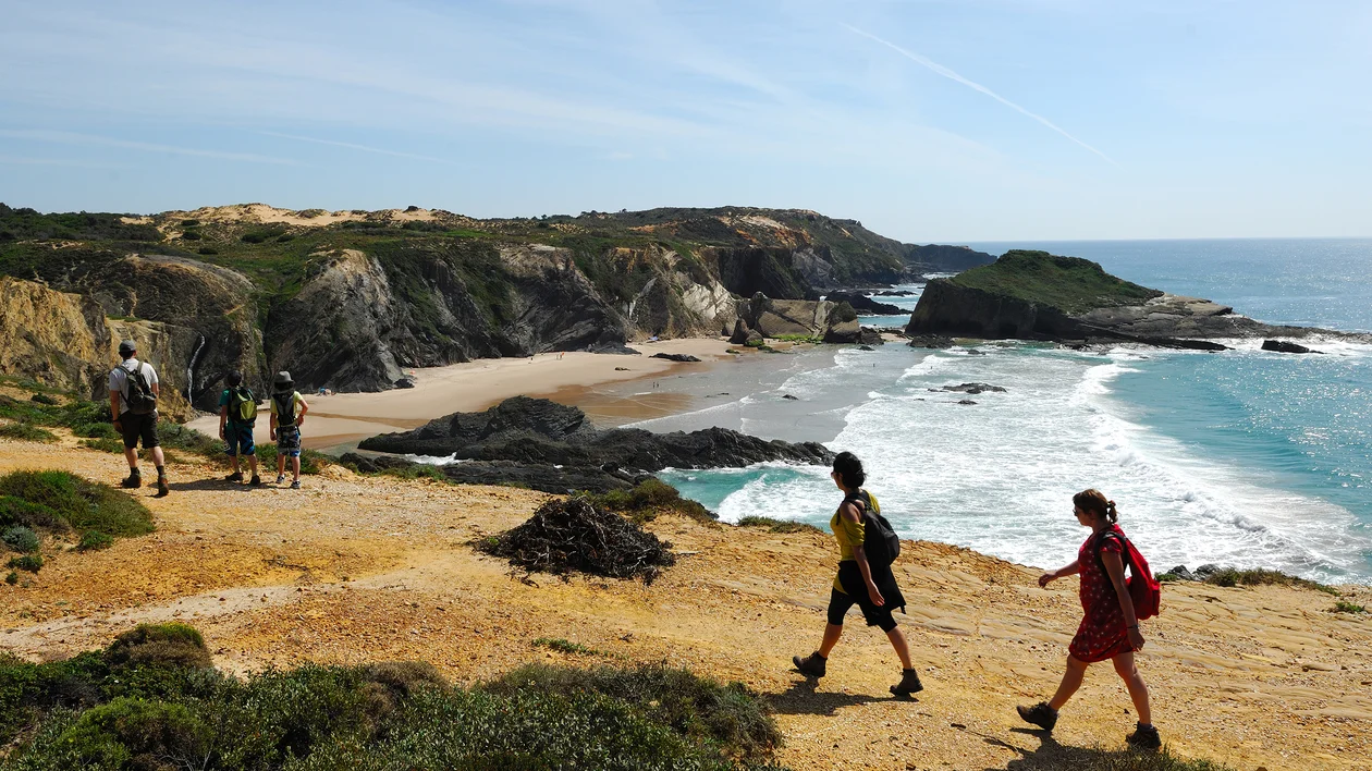 Hikers walking along a coastal cliff trail above a sandy beach and blue ocean waves.