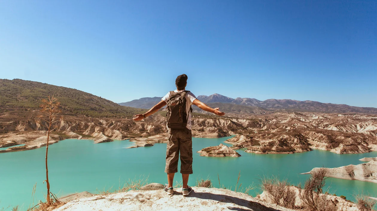 Person with outstretched arms standing on a cliff overlooking a turquoise lake and mountains.