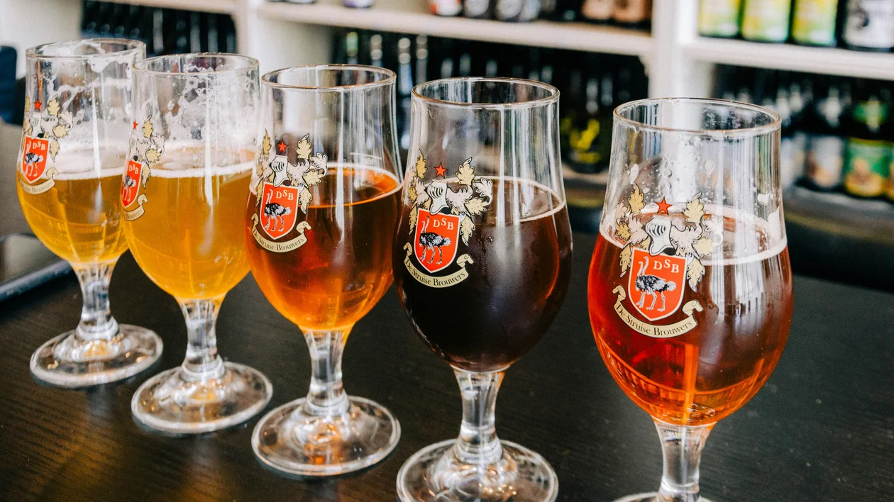 Five glasses of beer in varying shades on a dark table, bottles on shelves in background.